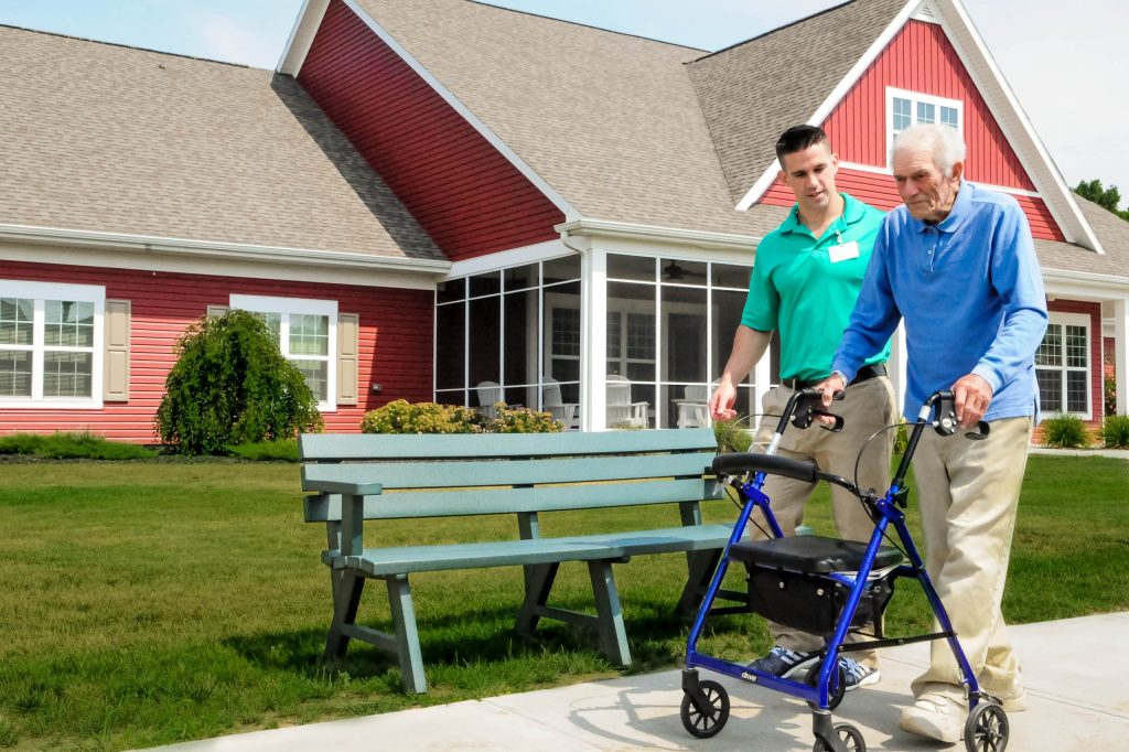 A therapist walks alongside a resident using a walker on a garden path